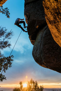 Side view of active climber in protective helmet hanging on mountain holding on to slope in warm light of sunset