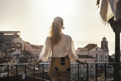 Rear view of young woman looking at city buildings against sky during sunset