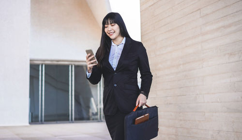 Smiling businesswoman using mobile phone while standing against wall