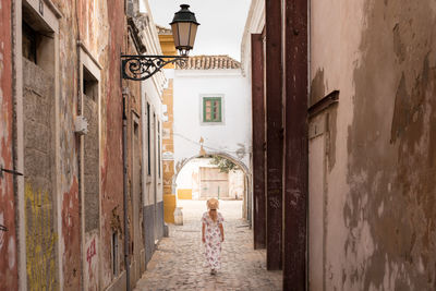 Back view of anonymous young female traveler in casual summer clothes walking along narrow paved street in algarve, portugal