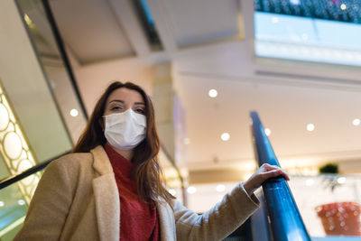 Low angle portrait of woman wearing mask sanding on escalator at shopping mall