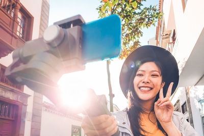 Smiling young woman gesturing peace sign while taking selfie in city during sunny day