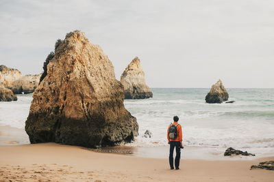 Rear view of man standing by rock at beach against sky