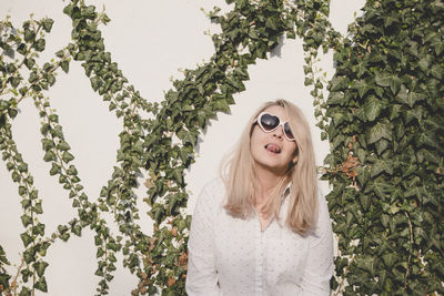 Young woman wearing heart shape sunglasses sticking out tongue while standing against wall