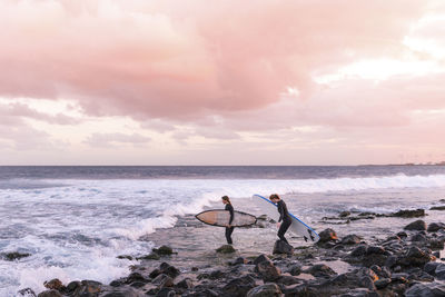 Female friends carrying surfboards on shore at beach against cloudy sky
