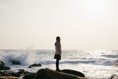 Side view of young woman standing on rock against splashing sea waves