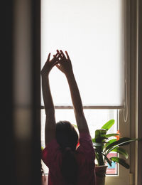 Rear view of girl with arms raised looking through window at home