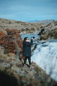 Woman photographing through camera while standing on rock against sky