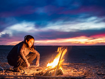 Woman crouching by campfire during sunset