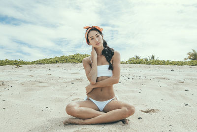 Portrait of woman in bikini sitting at beach against cloudy sky