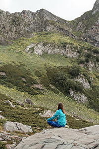 Rear view of meditating young woman in blue sitting on stone in mountain valley, consciousness