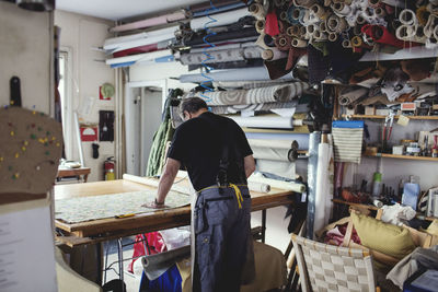 Rear view of owner working on fabric by shelves at workshop