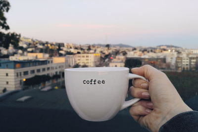 Cropped image of person holding coffee cup with text on it against city during sunset