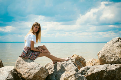 Young woman sitting on rocks by sea against sky