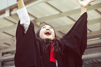 Cheerful young woman in graduation gown with arms raised 