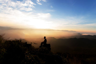 Silhouette man sitting on rock by landscape against sky during sunset