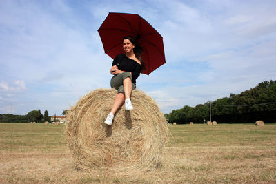 Young woman on a hay ball in a plowed field in tuscany in summer under the umbrella enjoying the sun