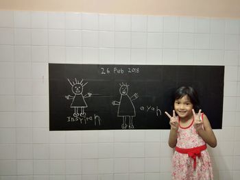 Portrait of cute smiling girl gesturing peace sign against chalk drawing on wall