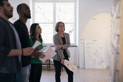 Multi-ethnic male and female engineers holding documents while reading notes on wall in office