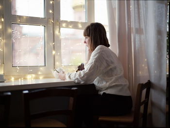 Side view of young woman using mobile phone while sitting by window at home