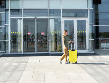 Young woman traveler carrying yellow suitcase next to entrance airport outside tourism 