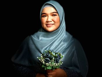 Portrait of smiling woman wearing hijab holding flowers against black background