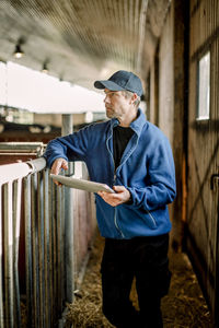 Farmer wearing cap standing with digital tablet by railing at cattle farm
