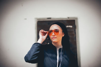 Portrait of young woman wearing sunglasses standing against wall