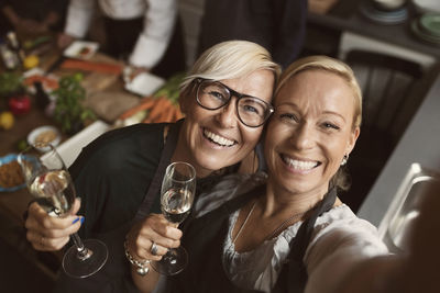 Cheerful mature women holding champagne flutes while taking selfie in kitchen