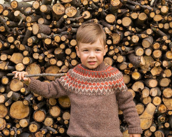 A boy in a lopapeis sweater holds a stick in his hands and stands against the background of firewood