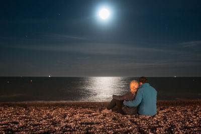 Couple sitting on shore against sky at night