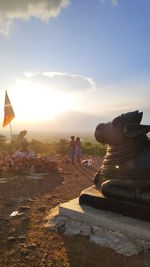 Man photographing sculpture against sky during sunset
