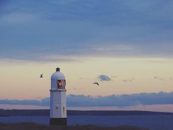 Seagulls flying past a lighthouse by sea against sky during sunset