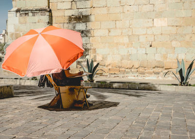 Low section of artist with orange parasol sitting on street against wall