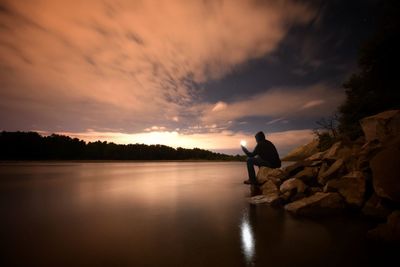 Optical illusion of man holding sun while sitting on rock by lake against sky during sunset