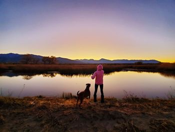 Rear view of woman standing with dog by lake during sunset