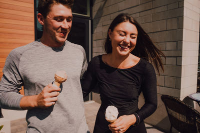 Crop adult man and positive fit woman with closed eyes in casual clothes having fun and laughing while walking along street and eating yummy ice cream