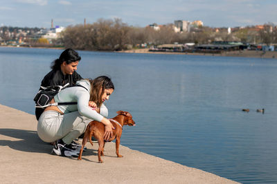 Young girl and her mother on relaxing vacation by the lake with their cute miniature pinscher.