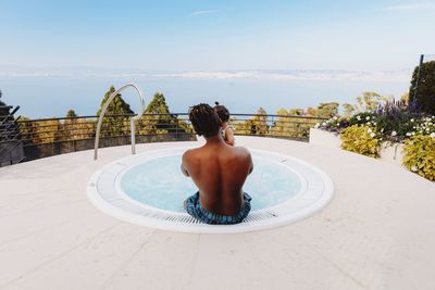 Rear view of woman sitting by swimming pool against sky