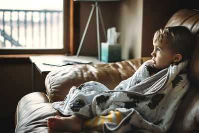 Thoughtful boy wrapped in blanket relaxing on sofa at home