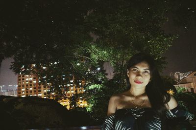 Portrait of young woman standing against trees at night