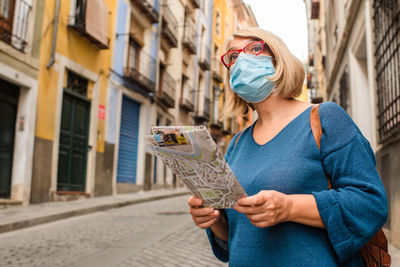 Low angle of female traveler in medical mask for coronavirus prevention holding touristic map and checking route while standing on narrow paved street with old buildings in cuenca town in spain