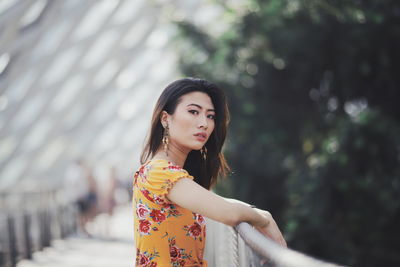 Portrait of beautiful young woman standing by railing against trees