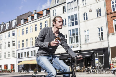Young man holding burger while riding bicycle on city street