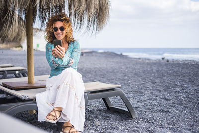 Full length of young woman using mobile phone while sitting at beach