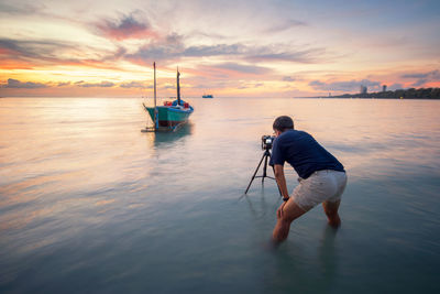 Man photographing boat while standing in sea against sky during sunset