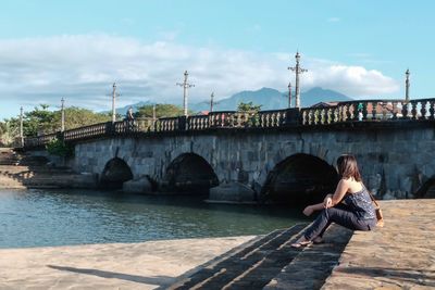 Woman sitting on bridge over river against sky in city