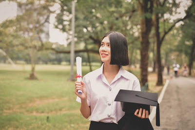 Young woman holding mortarboard and graduation gown on street