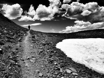 Rear view of hiker walking on dirt road against cloudy sky