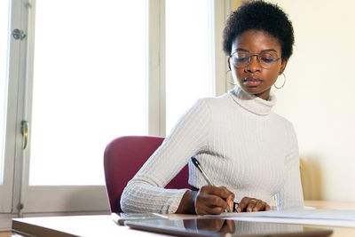 Black female physician writing information on paper sheet while preparing medical report at table in office of modern clinic